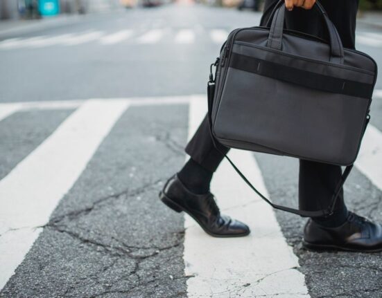 A woman in a stylish yet comfortable flat shoe walks purposefully across a city street, carrying a laptop bag, depicting a daily commute.