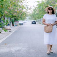 A woman in a flowing, light blue linen midi dress walks confidently down a sunlit European city street, embodying comfortable women's summer outfits for humid weather.