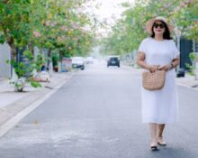 A woman in a flowing, light blue linen midi dress walks confidently down a sunlit European city street, embodying comfortable women's summer outfits for humid weather.