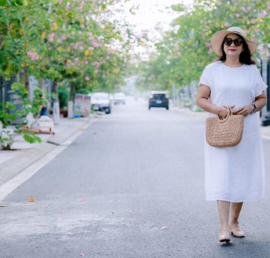 A woman in a flowing, light blue linen midi dress walks confidently down a sunlit European city street, embodying comfortable women's summer outfits for humid weather.