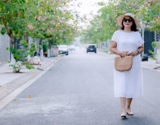 A woman in a flowing, light blue linen midi dress walks confidently down a sunlit European city street, embodying comfortable women's summer outfits for humid weather.