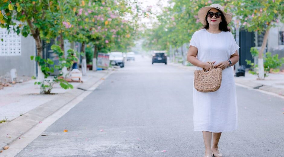 A woman in a flowing, light blue linen midi dress walks confidently down a sunlit European city street, embodying comfortable women's summer outfits for humid weather.