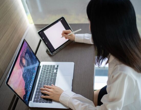 A woman smiling and working on a laptop at her home office desk, wearing a stylish yet comfortable knit sweater and tailored trousers, reflecting easy workwear outfits for hybrid offices.